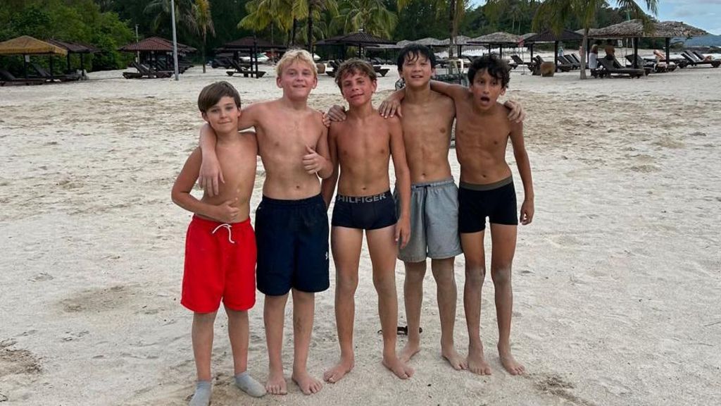 Five boys stand on a beach. Several of them have their arms around each other's shoulders. All are wearing swimming trunks.