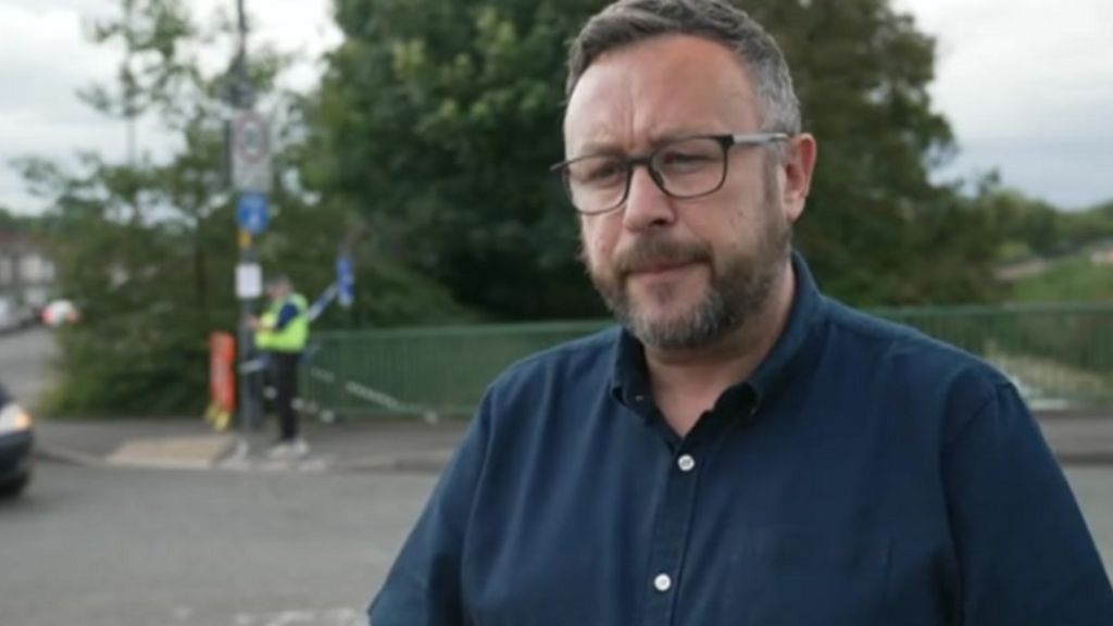 A man with a beard and glasses, wearing a blue shirt looks at the camera. Behind him is a canal bridge and a policeman standing next to a cordon