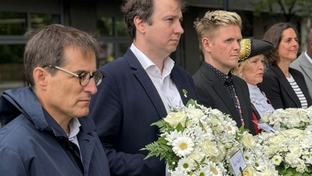 A group of people in formal attire stand outdoors holding wreaths and bouquets of flowers, including white and red arrangements with ribbon, during a ceremonial gathering.