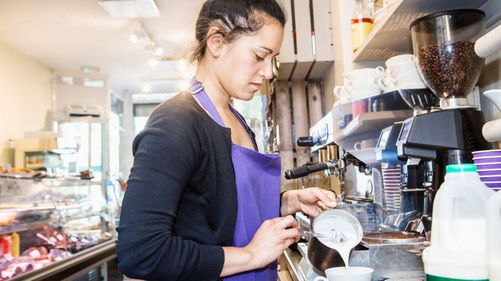 A woman wearing a purple apron works in a cafe. She is pouring milk into a mug next to a coffee machine
