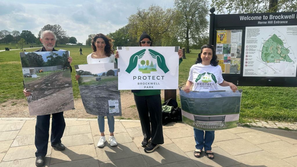 A group of four members of the organisation 'Protect Brockwell Park' standing in a line holding large posters of the park, in the park, with the grass looking muddy and damaged. One of the women in the middle is holding a Protect Brockwell Park Poster. 