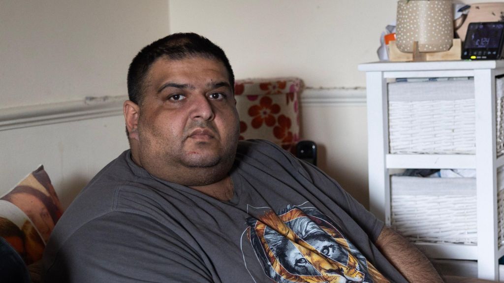 Amit Marwaha sits on a sofa holding a phone, wearing a T-shirt with an image of a tiger printed on it. Behind him is a white storage unit with baskets, flowers, and a clock.