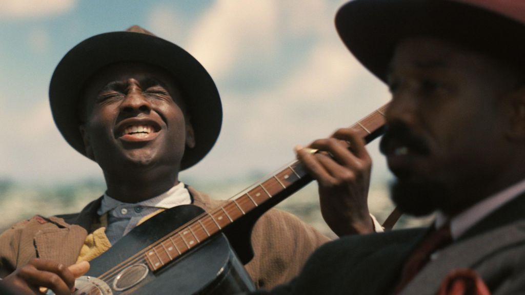 A still from Sinners showing Miles Caton as Sammie playing a guitar and singing and Michael B Jordan in the foreground looking ahead as he drives a car