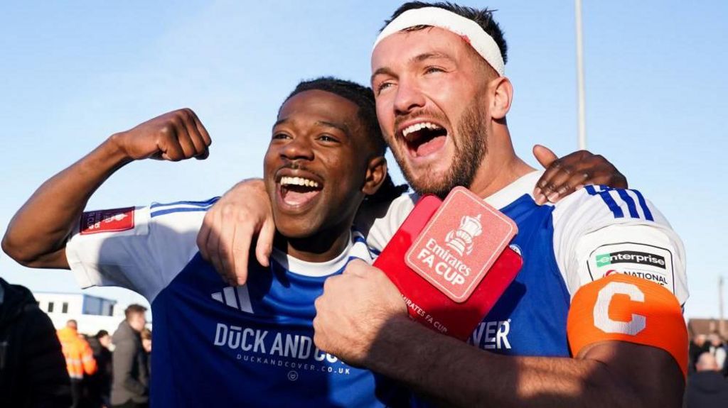 Macclesfield FC goal scorers Isaac Buckley-Ricketts and Paul Dawson celebrate following the Emirates FA Cup third round match at the Leasing.com Stadium, Macclesfield. They are wearing Macclesfield FC kits and have big smiles, with Buckley-Ricketts raising his arm in the air in celebration. Dawson, with a captain armband, holds a card with the Emirates FA Cup logo on it.
