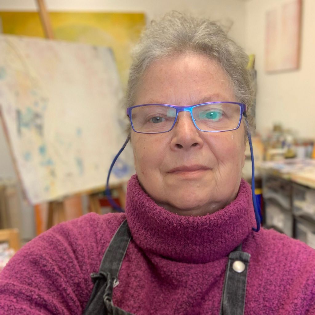 Denise takes a selfie in an art studio as she wears a dark pink turtle-neck jumper with an apron on top.