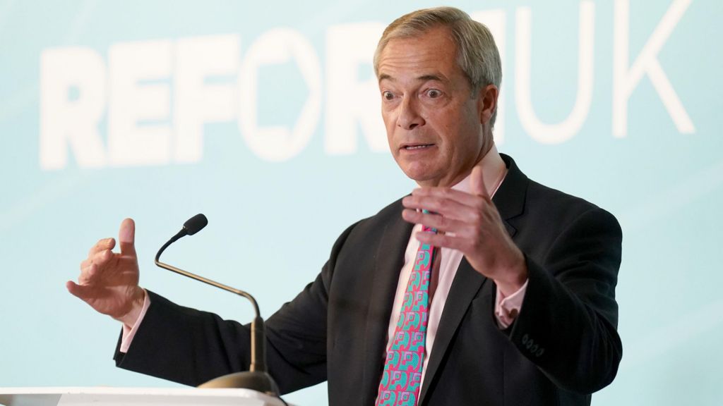 Nigel Farage, a middle aged man with greying hair in a side parting, is standing in front of a light blue background making a speech. He is wearing a dark suit, pink shirt and coloured patterned tie.