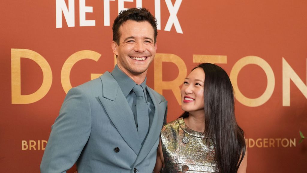 Luke Thompson smiles at the camera in a blue suit and tie and is stood beside Yerin Ha who wears a metallic dress and has long dark hair. They are stood against an orange screening backdrop