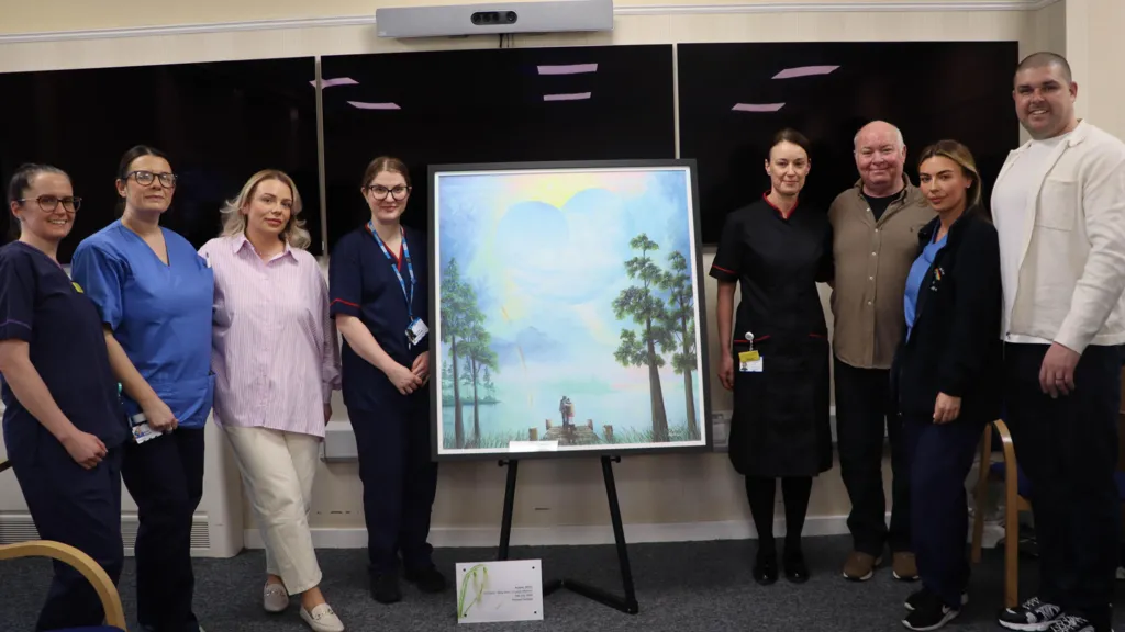Eight people standing on either side of a piece of artwork, which is placed on an easel. Some of them are wearing blue medical uniform. The pastel artwork is of two figures arm-in-arm at the end of a wooden pier. The sky above the figures is blue, and is dotted with clouds including a heart shaped one. The scene is bordered by tall trees.