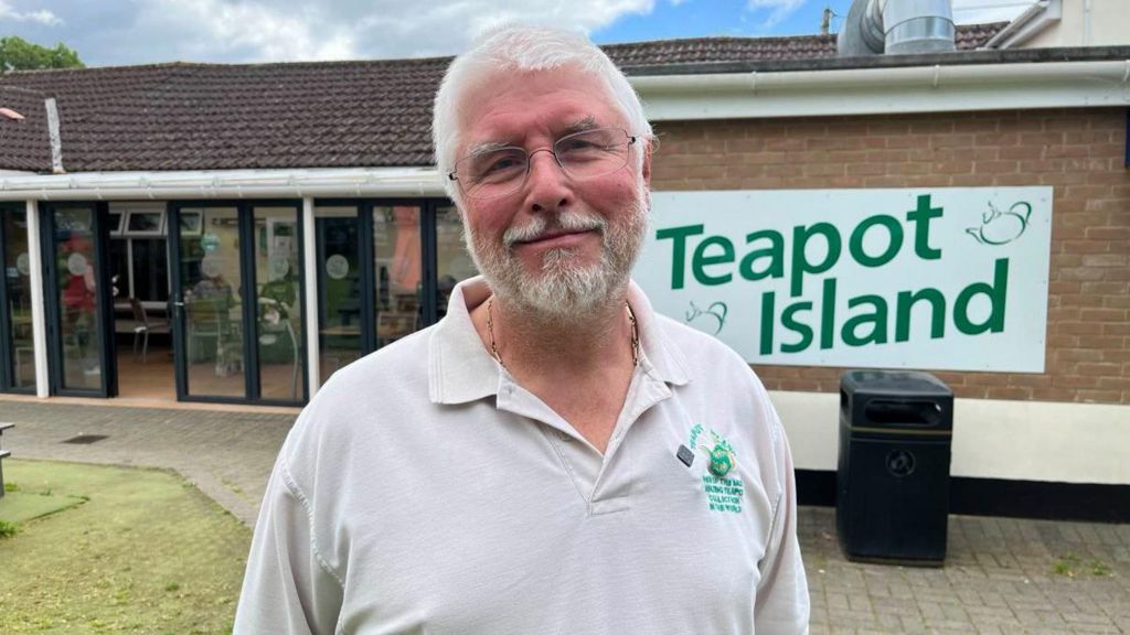 A man in his sixties with white hair and a white beard and glasses looks at the camera in front of his business premises, teapot island