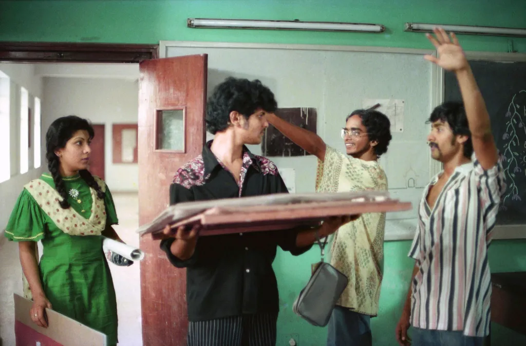 Four students - a woman in a green tunic along with three men, dressed in black, yellow and a striped shirt, respectively - stand in a classroom in a scene of the film. 