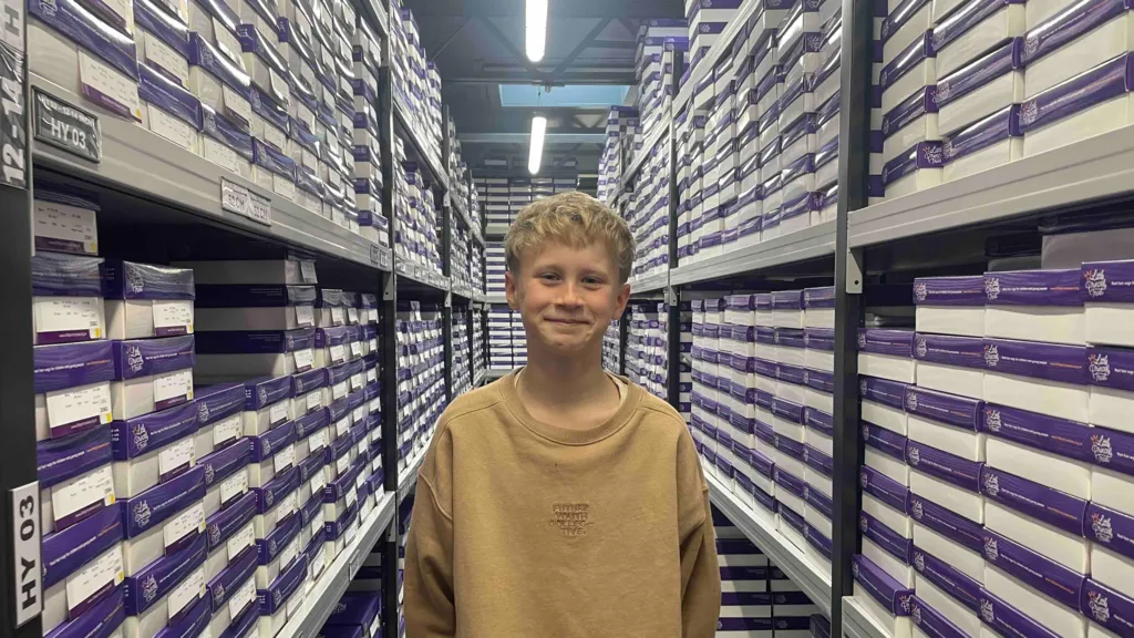 Jack, un garçon blond aux cheveux mi-longs, se tient dans un entrepôt, entouré de cartons.