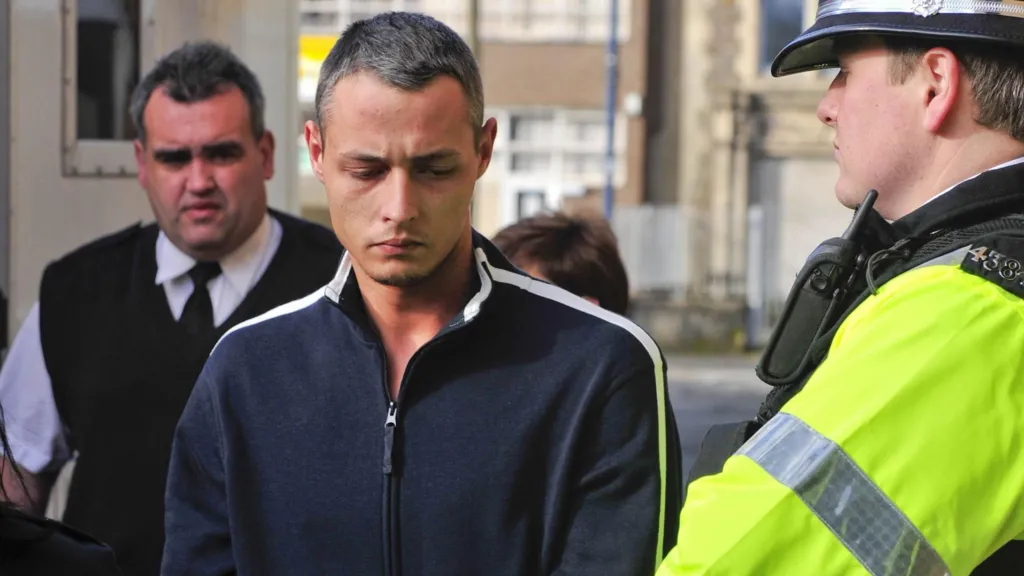 Paul Grabham, 25, arrives at Swansea Magistrates Court, where he is charged with the murder of his wife, Kirsty, 24. Paul is wearing a dark blue zip up top with white stripes down the shoulders and sleeves and is staring at the ground as he walks. A prison van is visible over his right shoulder and a prison guard is walking behind him wearing a white shirt with a black tie and a gilet style jumper over the top. To Paul's immediate left is a police officer in full uniform looking at him
