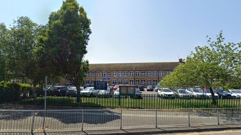 The entrance of Pensby High School with a blue sign on railings and the brown long building in the background, behind cars parked in the car park. 