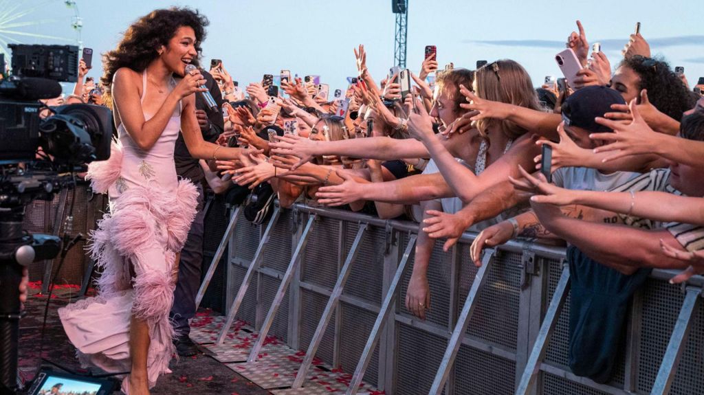 Olivia Dean walks along a crowd barrier - touching the hands of dozens of fans, who are holding their harms out. She's singing into a microphone and wearing a long pink dress. 