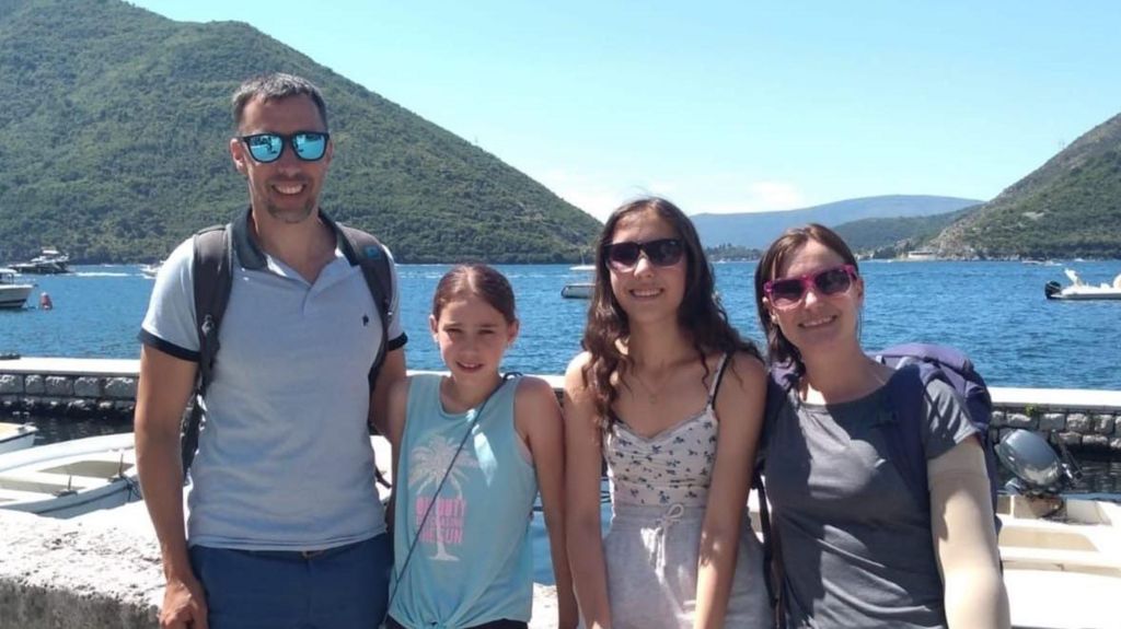 Sqn Ldr Mark Long, wearing sunglasses, a light blue polo shirt and a backpack, stands with his wife and daughters in front of a blue sea, blue sky and green hills 