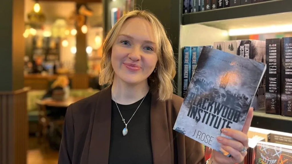 A woman with short blonde hair is wearing a black top and a brown blazer. She is holding a book that says 'Blackwood Institute'. 