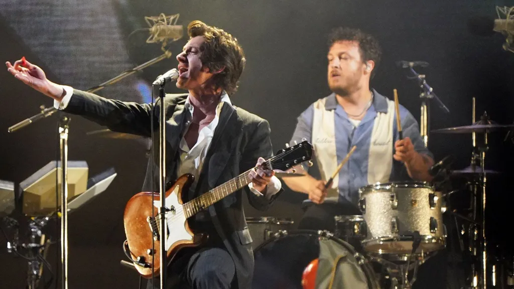 Frontman of Arctic Monkeys Alex Turner plays guitar on stage and drummer Matt Helders is seen closely behind him playing drums. Matt Helders is wearing a blue and white striped shirt and Alex Turner is wearing a black suit jacket and white shirt 