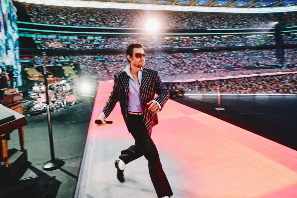Harry Styles strides onto the stage at Wembley Stadium, wearing a black and white pinstripe jacket and a wide-collared blue shirt. Spotlights illuminate the audience, and the singer is wearing aviator sunglasses.