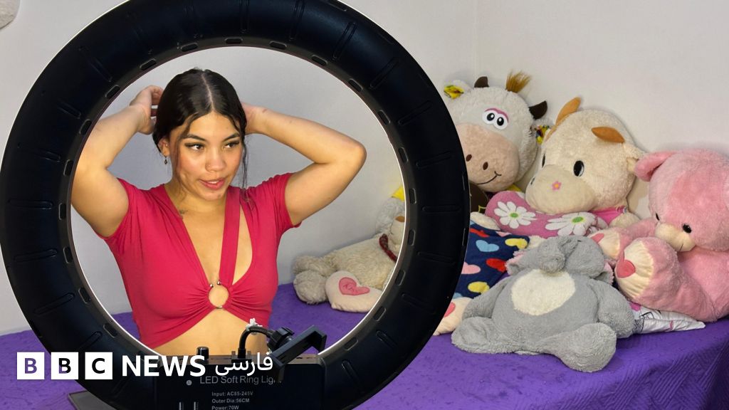 زن جوانی با تاپ کوتاه قرمز، روی تختی روبروی یک چراغ حلقه‌ای بزرگ نشسته است. موهای بلند و تیره‌اش را از پشت بسته است. کنار او روی تخت، عروسک‌های پارچه‌ای و اسباب‌بازی‌هایی قرار دارد