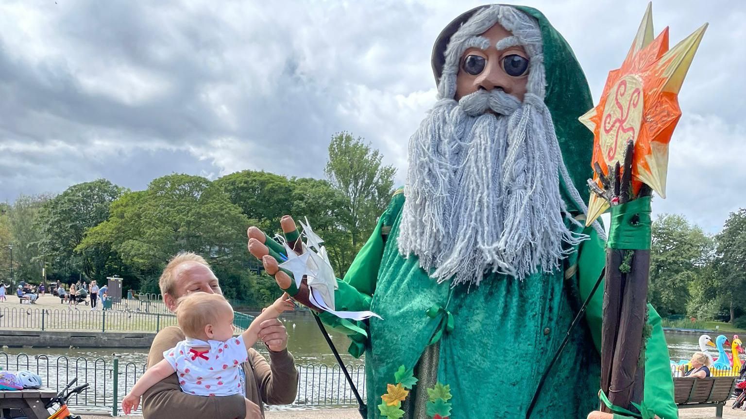 A man and young baby reach out to touch a giant green wizard puppet with long white hair and beard wearing a green cloak at an event by water