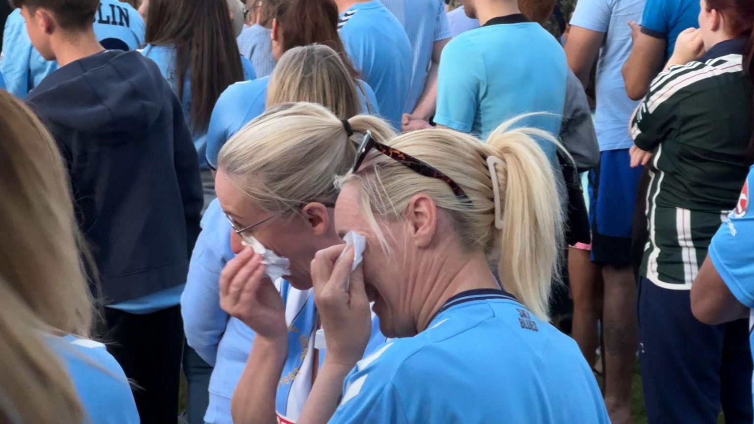 A crowd of people, many wearing sky blue colours, with two women wiping away tears in the foreground
