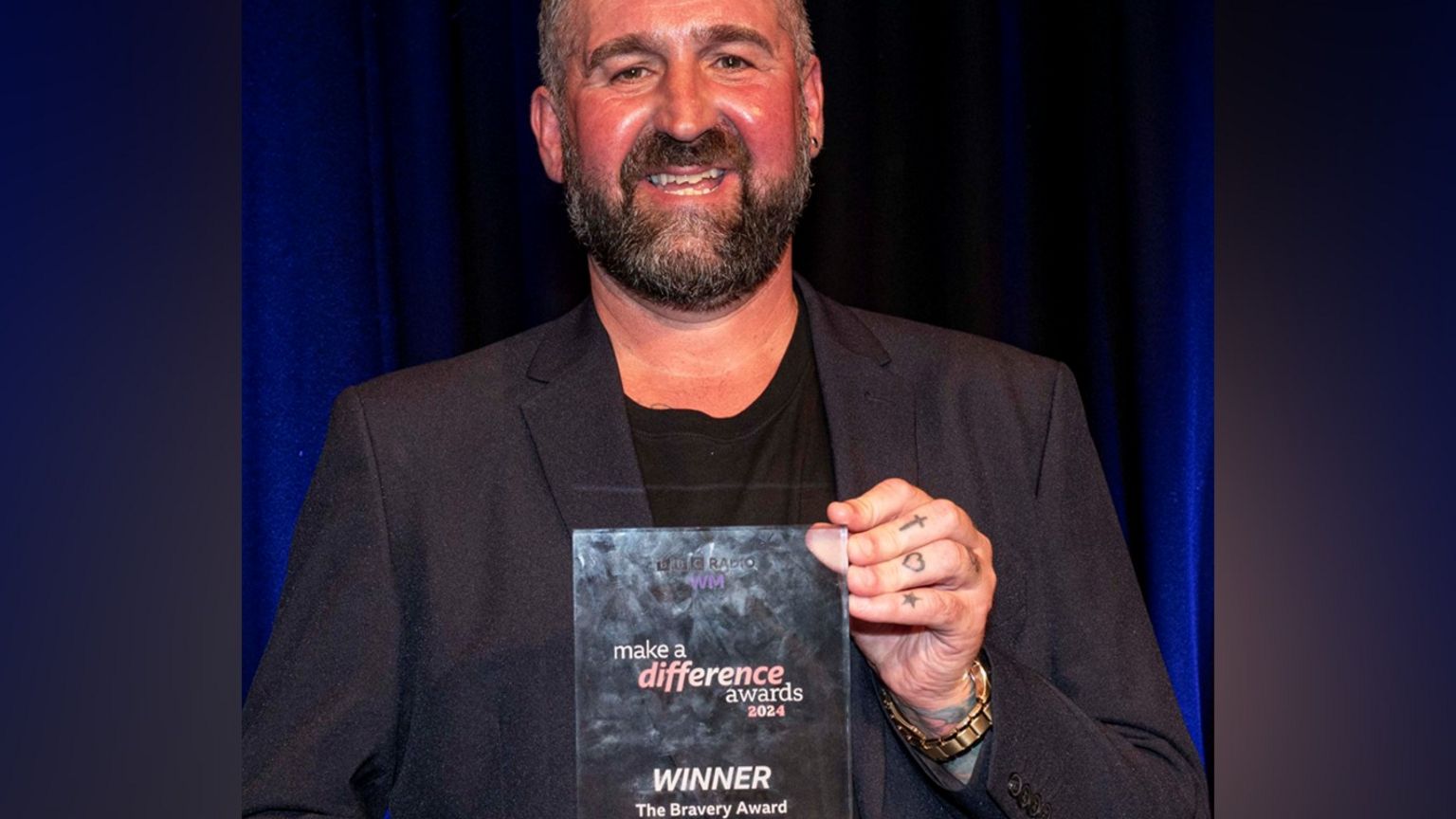 Steve Whitehouse is wearing a black suit and T-shirt underneath and is smiling holding his award and smiling in the photo. Steve has dark hair and a beard and is wearing a gold-coloured watch on his left hand and his fingers are also tattooed.


