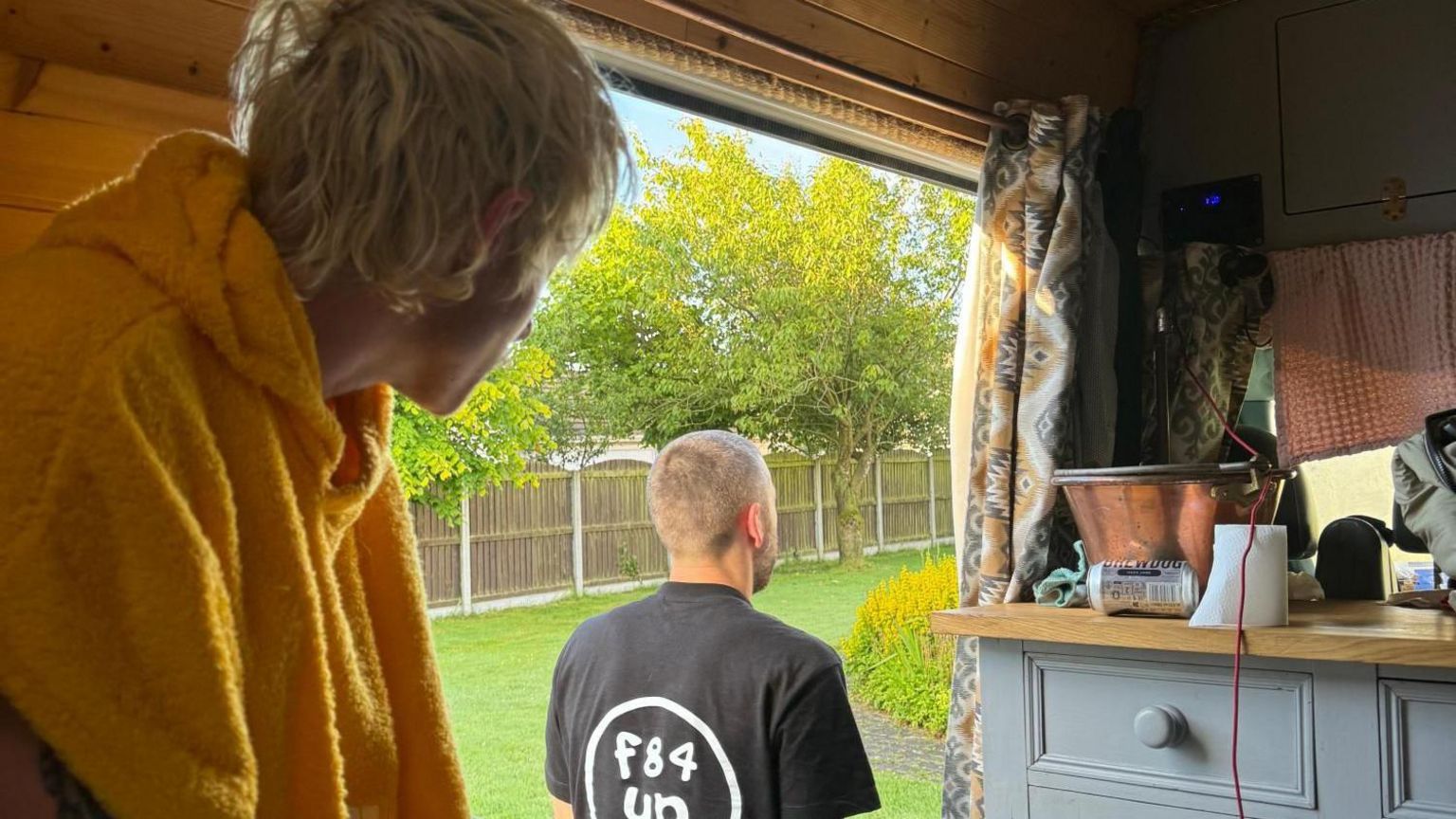 A young man with blonde hair and a yellow dressing gown looks out from the inside of a van