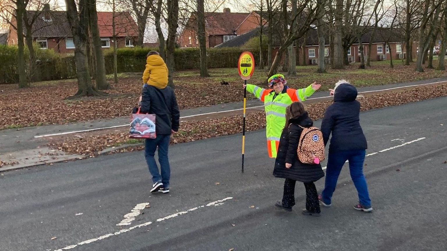 Lollipop lady holding lollipop sign while directing tow parents and their children across the roadtn