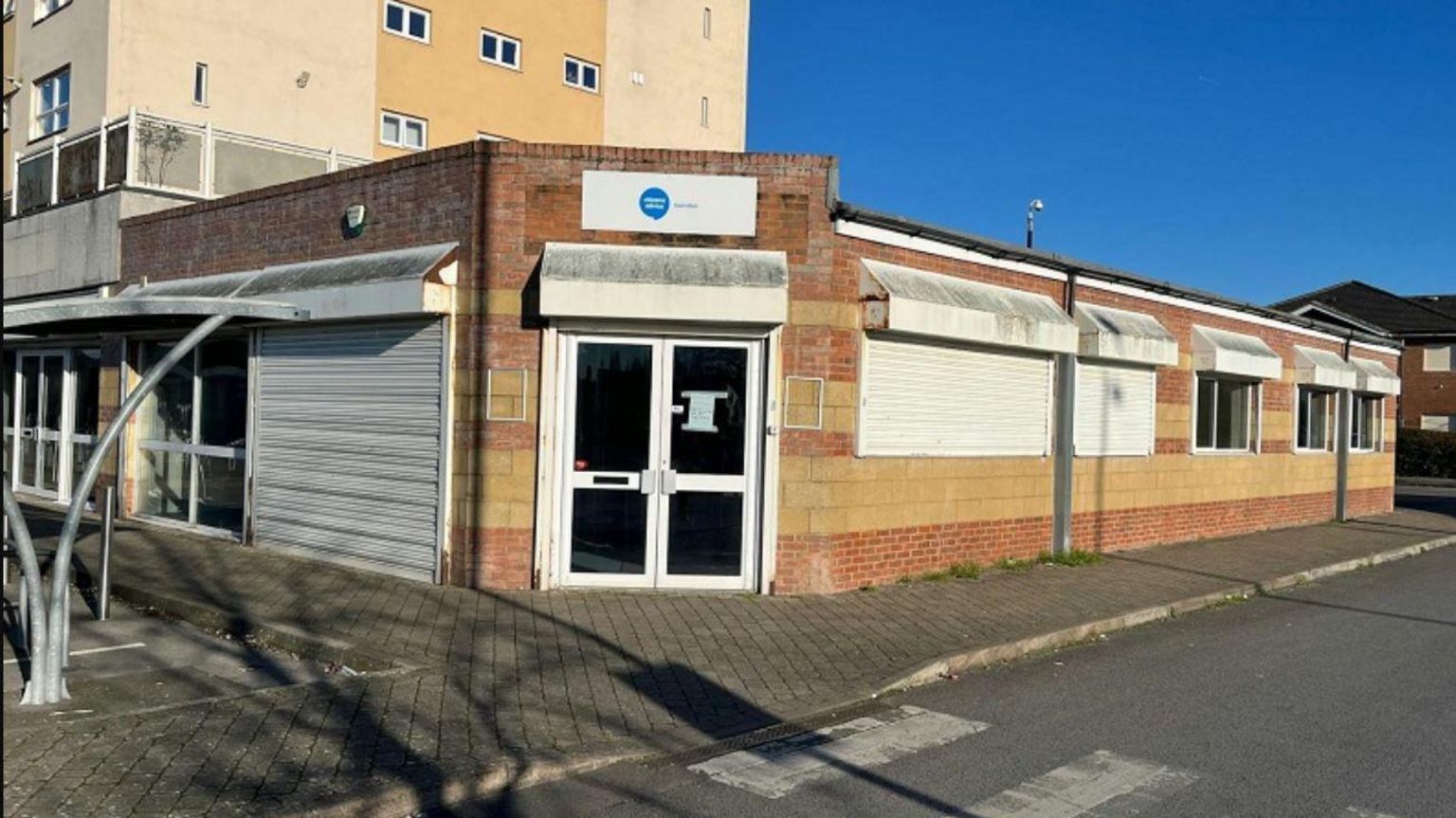 A shop with closed white shutters on the windows and the lights off inside. It is on the corner of Cavendish Square, with the front door facing the car park, and a tower block visible in the background.