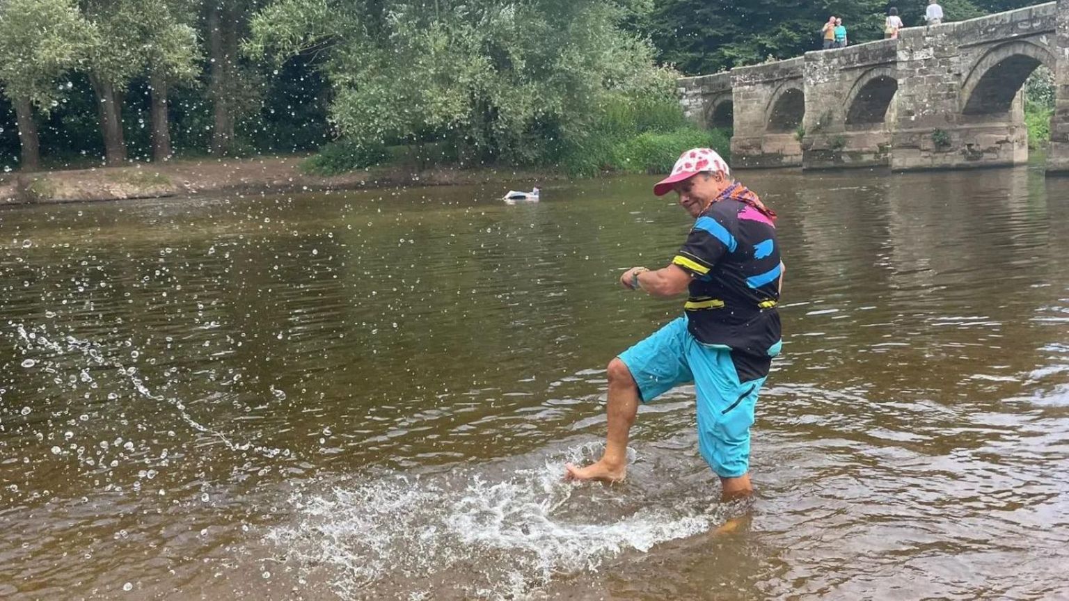 Timmy Mallet kicking water in a river. He is wearing a spotted baseball cap and bright blue trousers