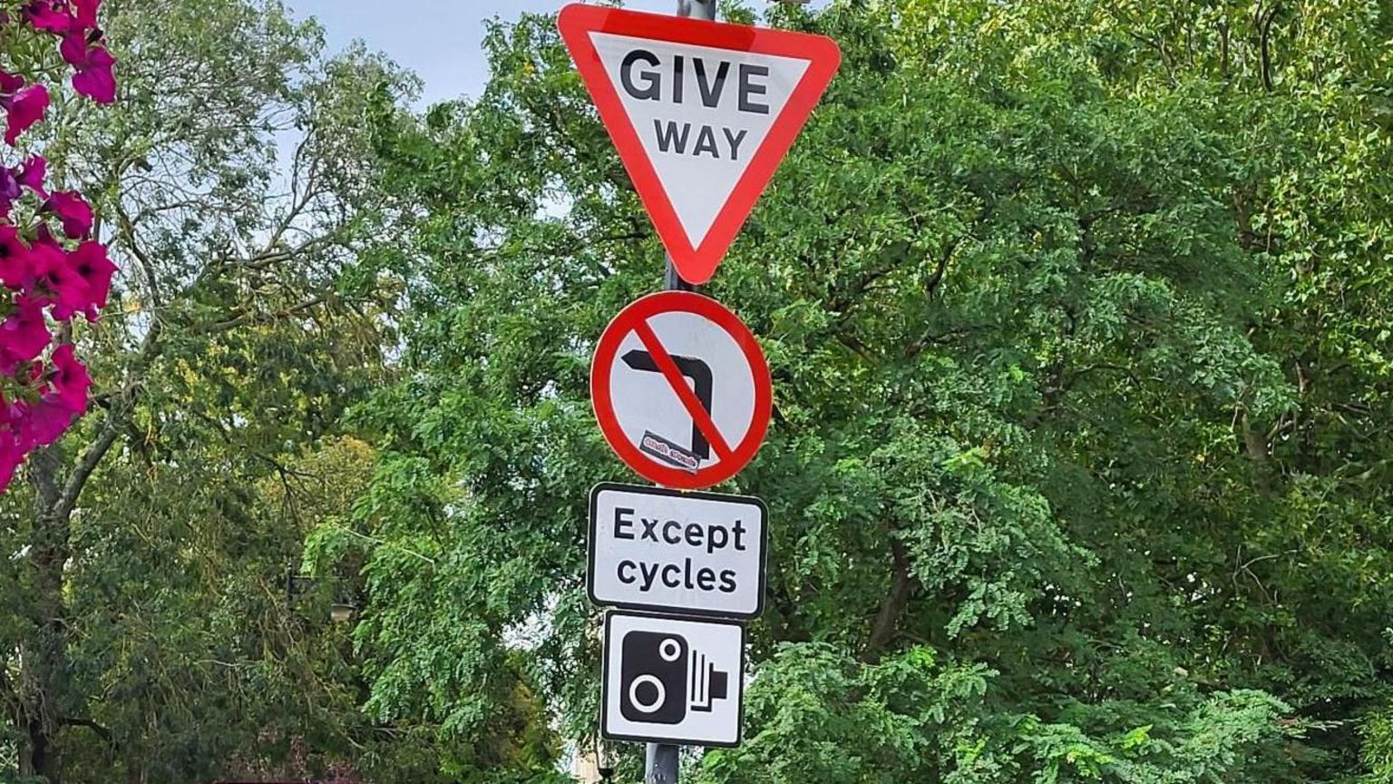 Three road signs on the Plain roundabout. At the top is a give way, then a no left turns, then one saying 'except cycles', and finally at the bottom is one of a camera.