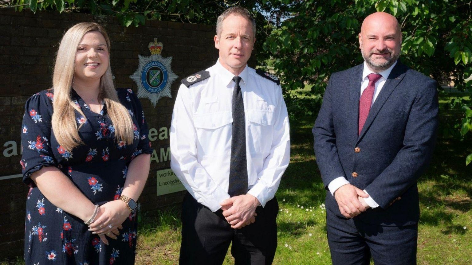 The three figures stand together in a garden area near a police headquarters.