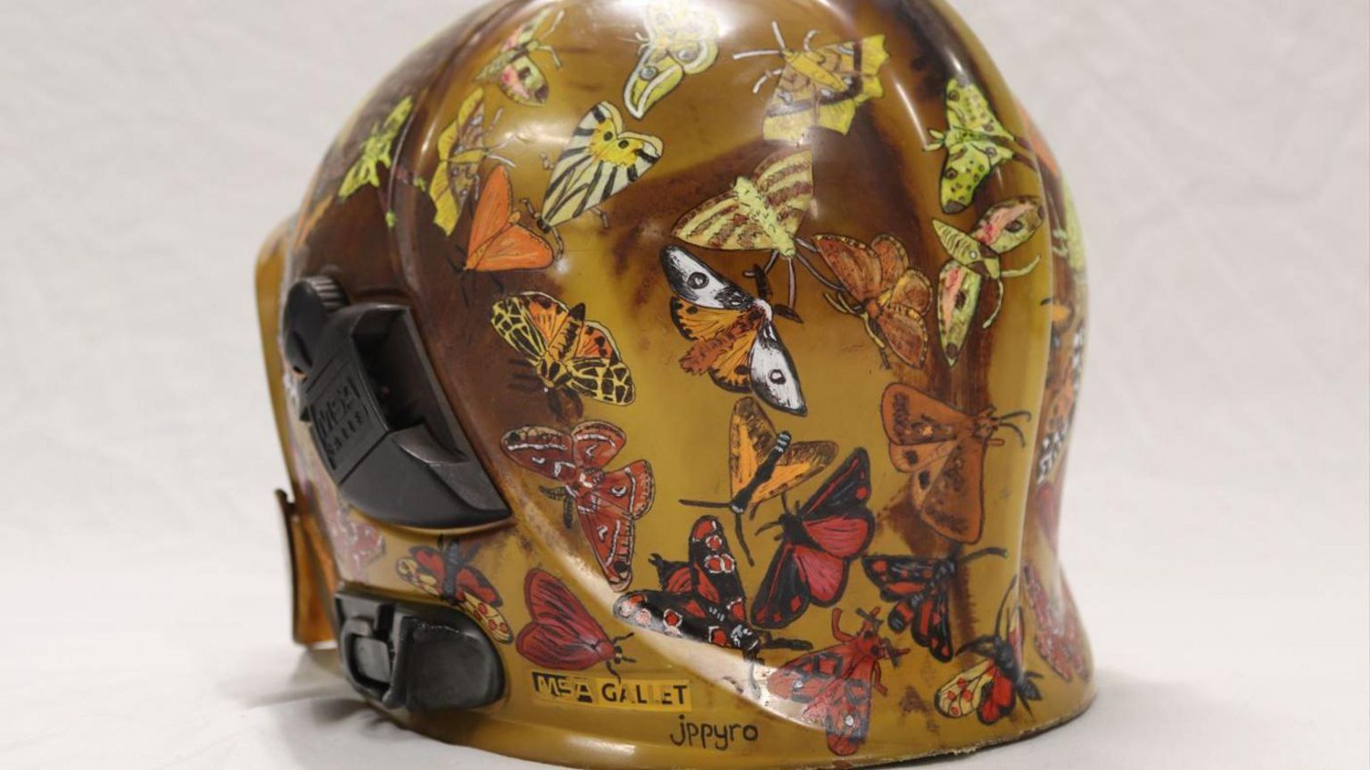 A firefighters helmet painted in shades of amber and brown with numerous colourful moths painted on top