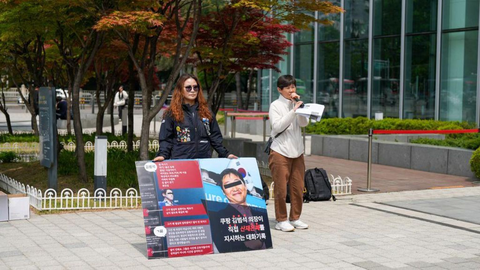 两名女子站在韩国一栋建筑物外。一名女子对着麦克风讲话，另一名女子则举着一张大型标语牌进行抗议。