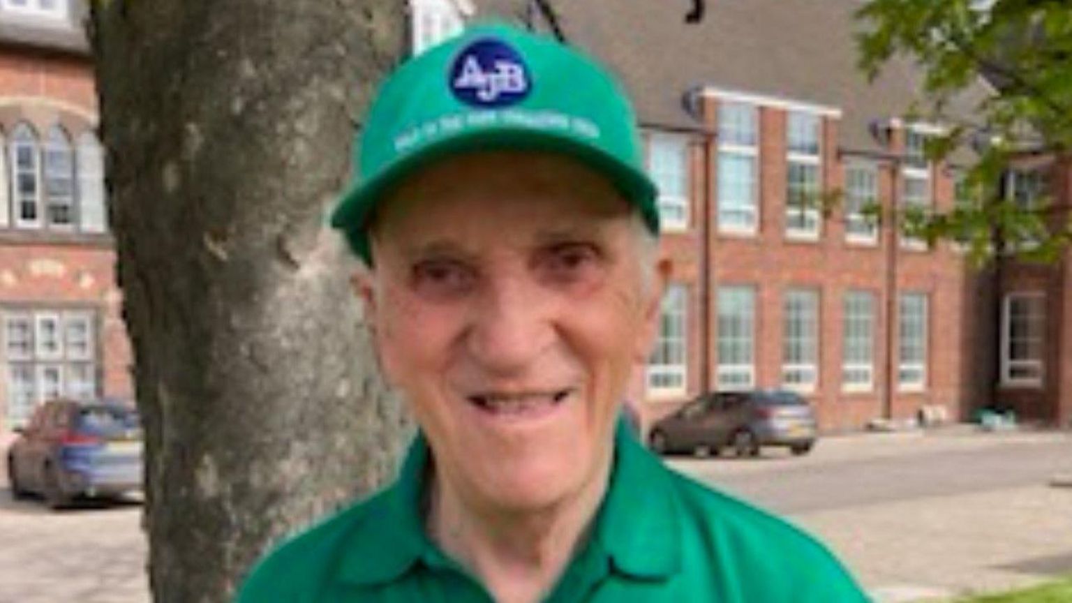 A man wearing a green baseball cap and green polo shirt, with the trunk of a tree and a red brick building in the background