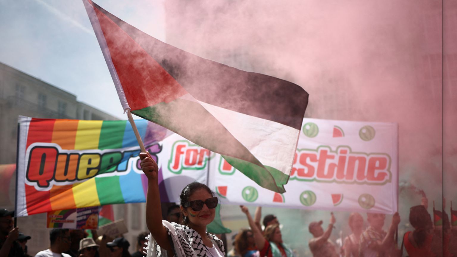 Woman waving a Palestinian flag with a large banner saying Queers for Palestine in the background.
