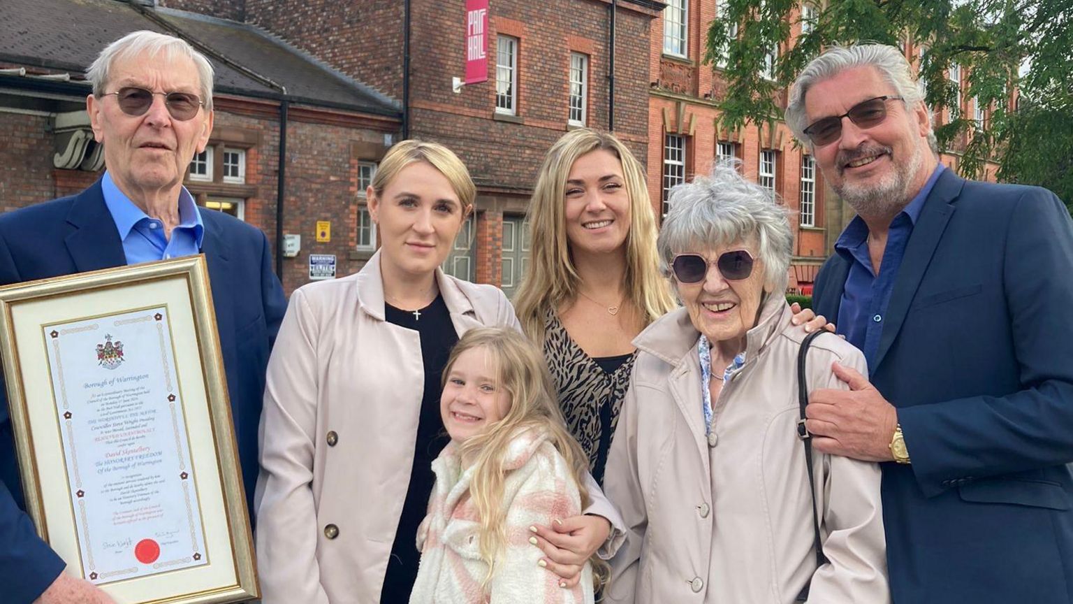 A smiling David Skentelbery and his son Gary pose with David's grey-haired wife Patricia, who wears dark glasses and wears a buff-coloured coat, and his two blond-haired granddaughters and blond-haired great-granddaughter