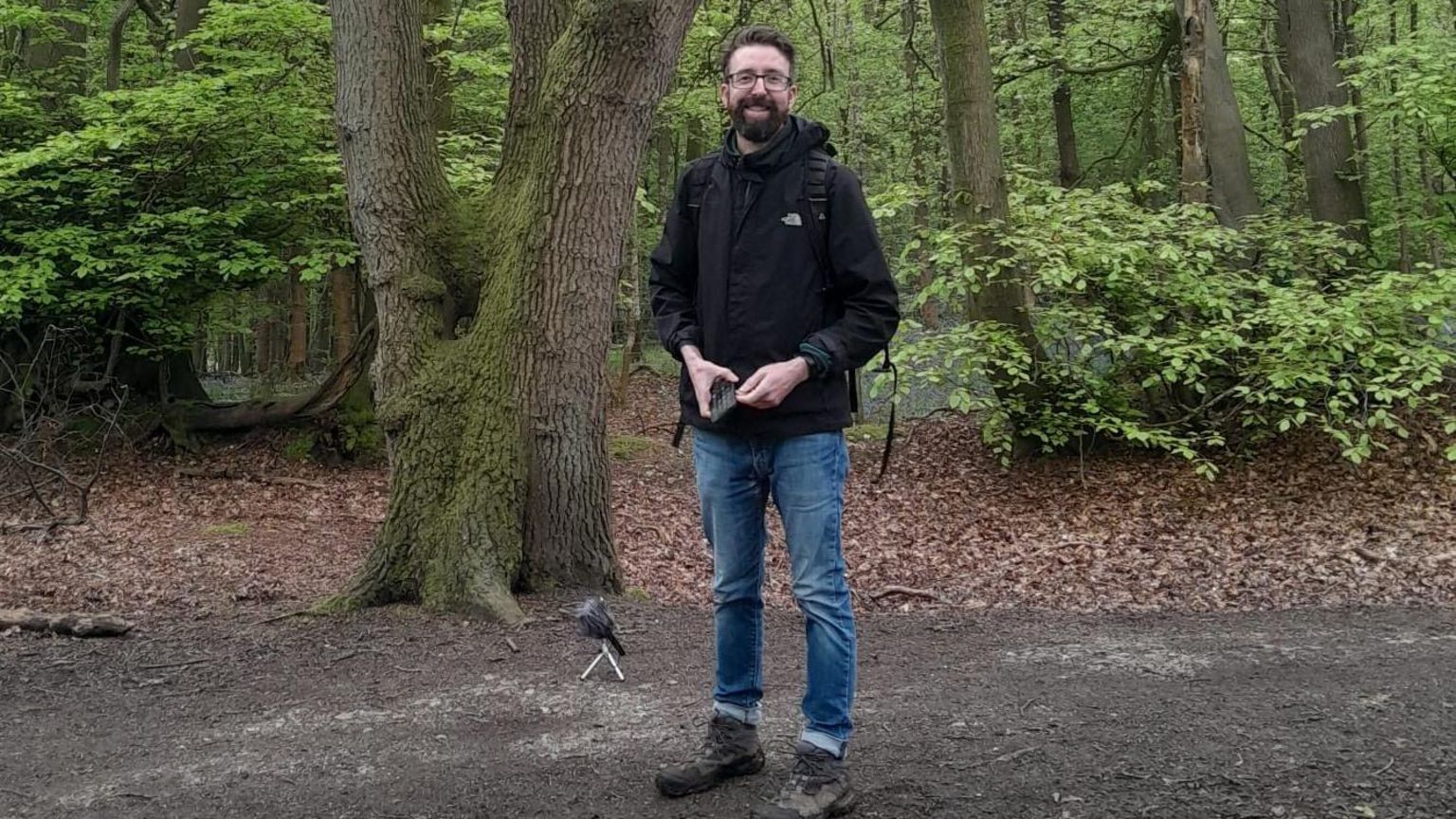 Paul Walker standing in Wendover Woods, by recording equipment, wearing blue jeans and a black jacket, looking straight at the camera and smiling