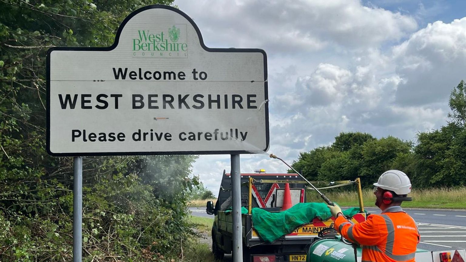 A council worker in an orange jacket cleaning a sign that says "Welcome to West Berkshire" with a pressure washer