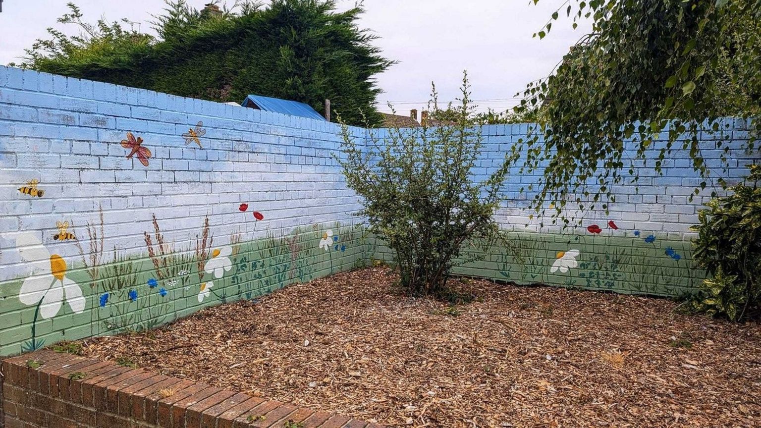 The light blue mural which has been painted onto a brick wall shows bees and dragonflies flying above grass and flowers