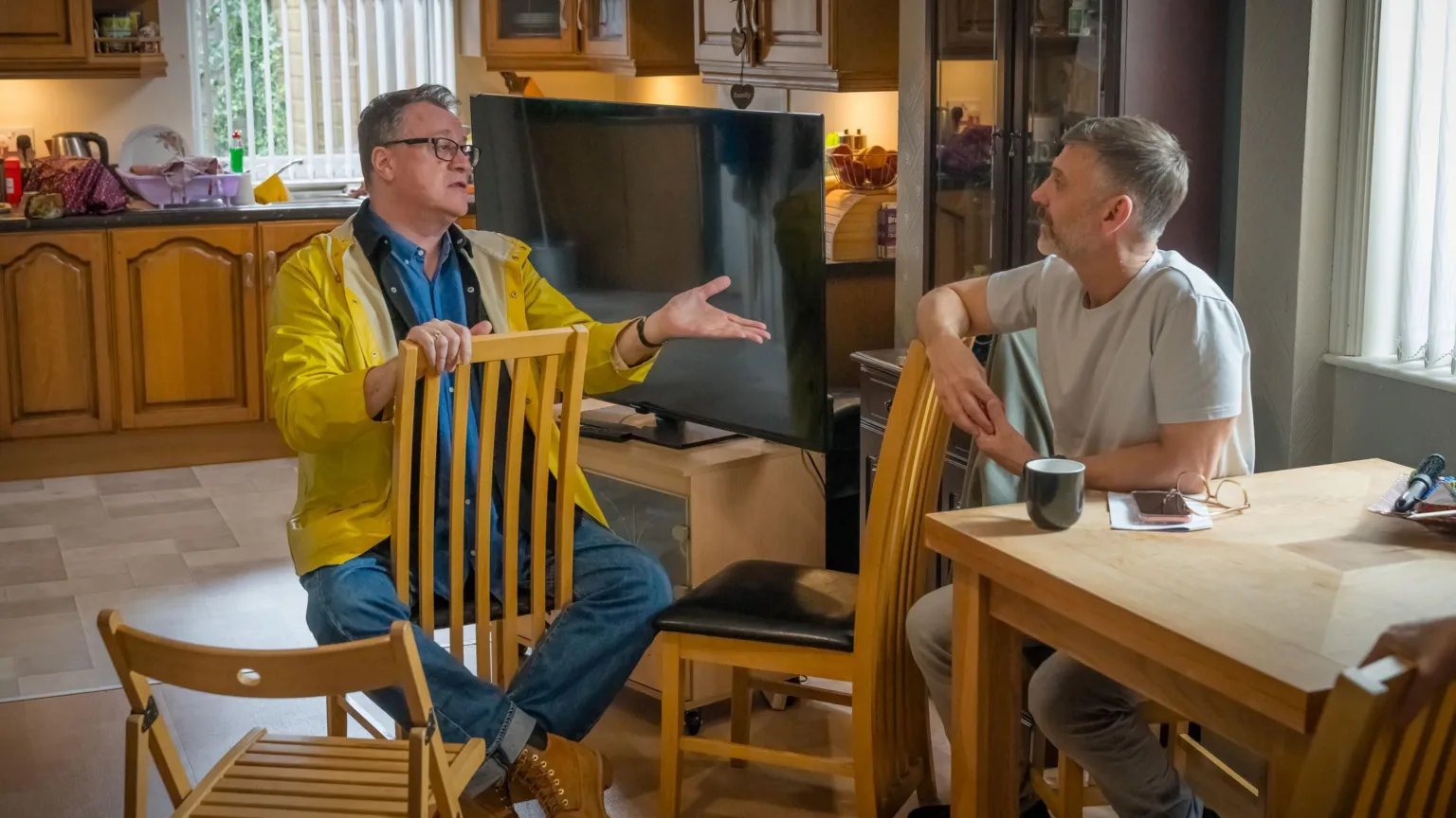 Channel 4 Russell T Davies and Peter Hoar are sat on a kitchen table discussing their work, the kitchen is a TV set. Russell is sitting backwards on the chair and gesticulating towards Peter who looks engaged.