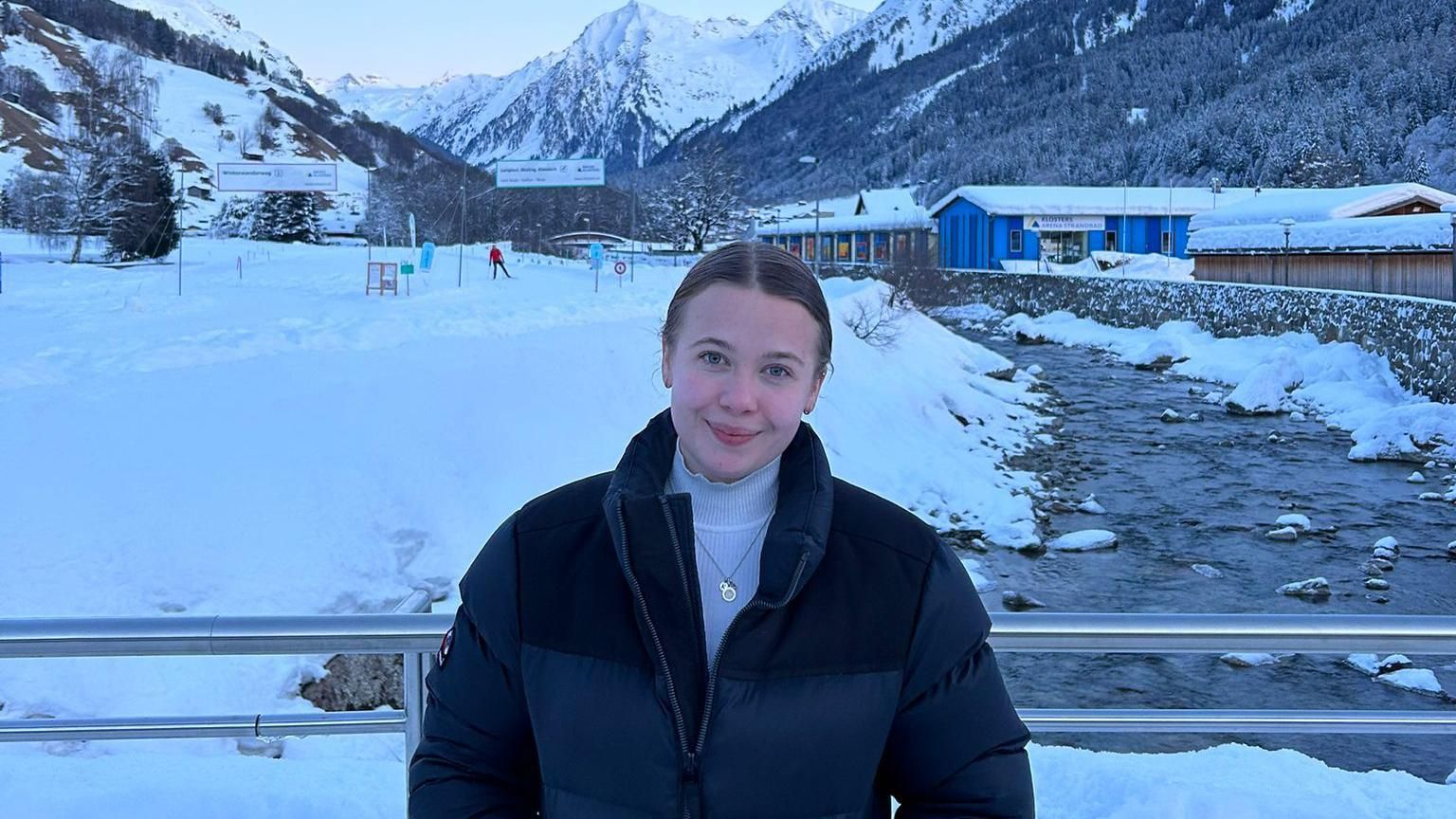 A young white woman wearing a warm navy blue jacket smiles at the camera in front of a snowy mountain scene