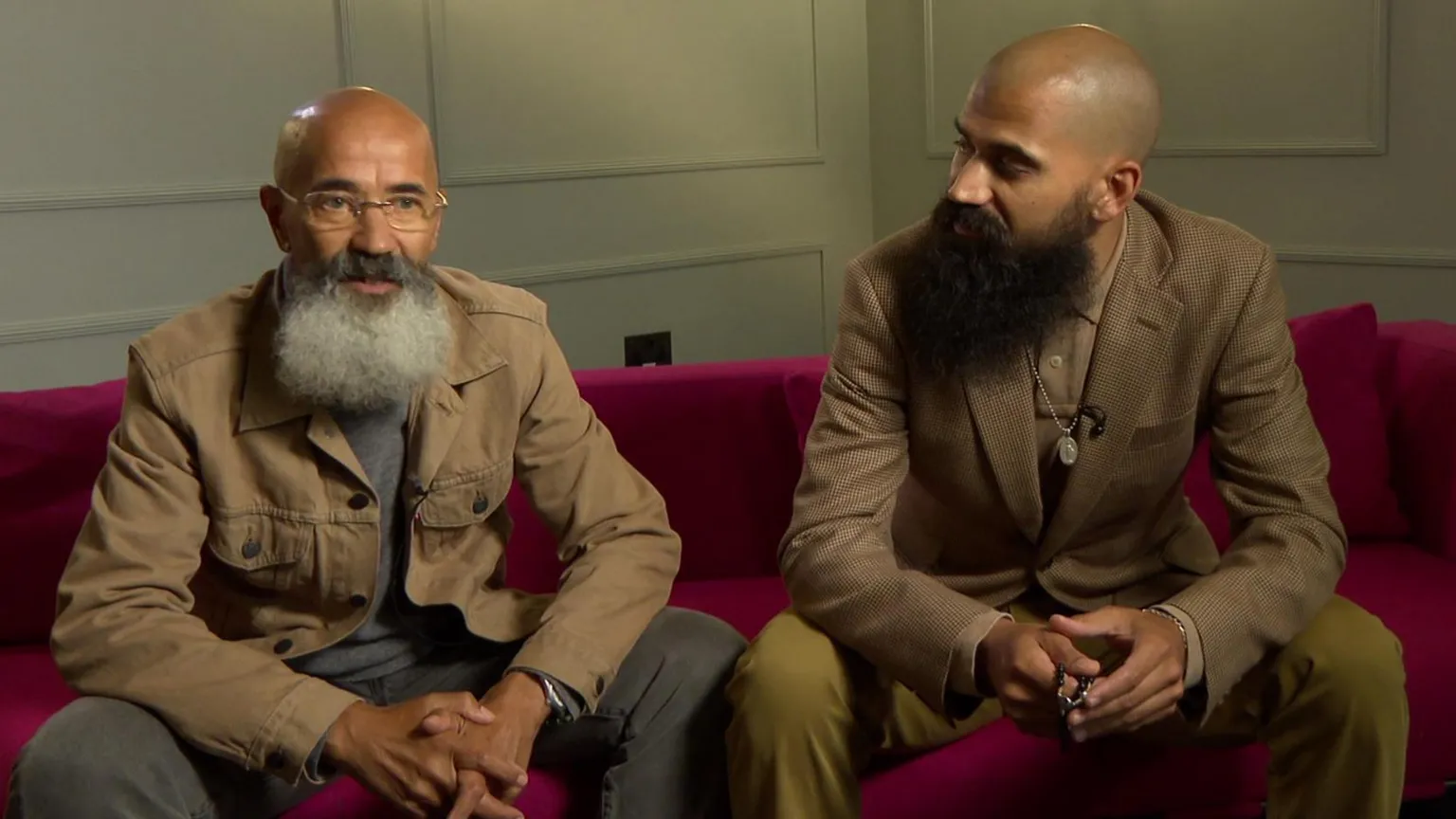 Two men with beards sitting on a red sofa indoors, one looking toward the other during an interview-style setting. They wear brown tan clothing.