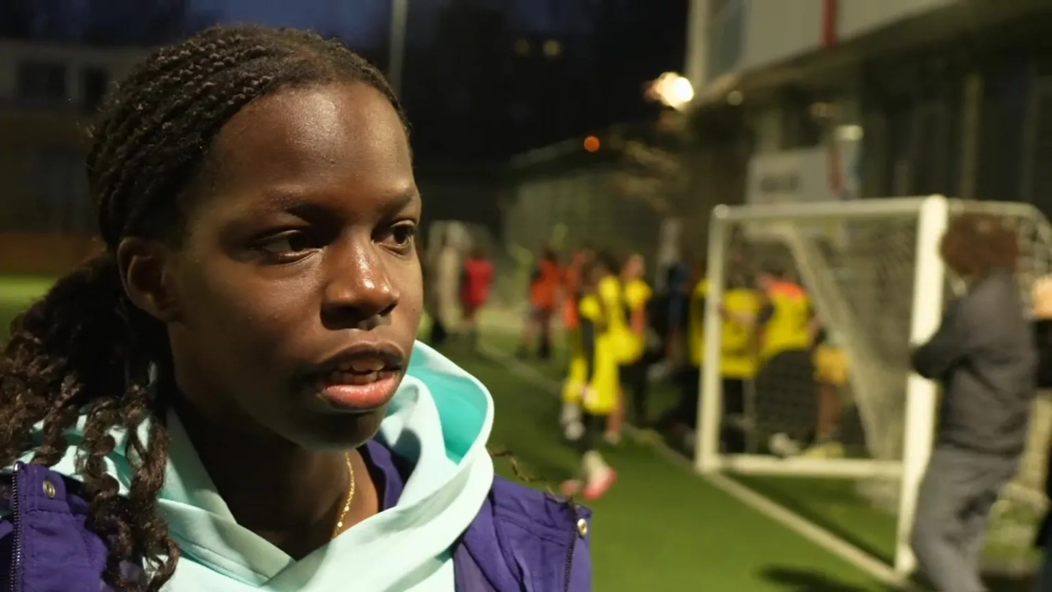 TV screenshot of Vivienne Lia, a footballer. She is standing by the edge of a football pitch on which a training session is taking place. It is dark and the pitch's lights are on. Vivienne is wearing a pale blue hoodie and a purple coat