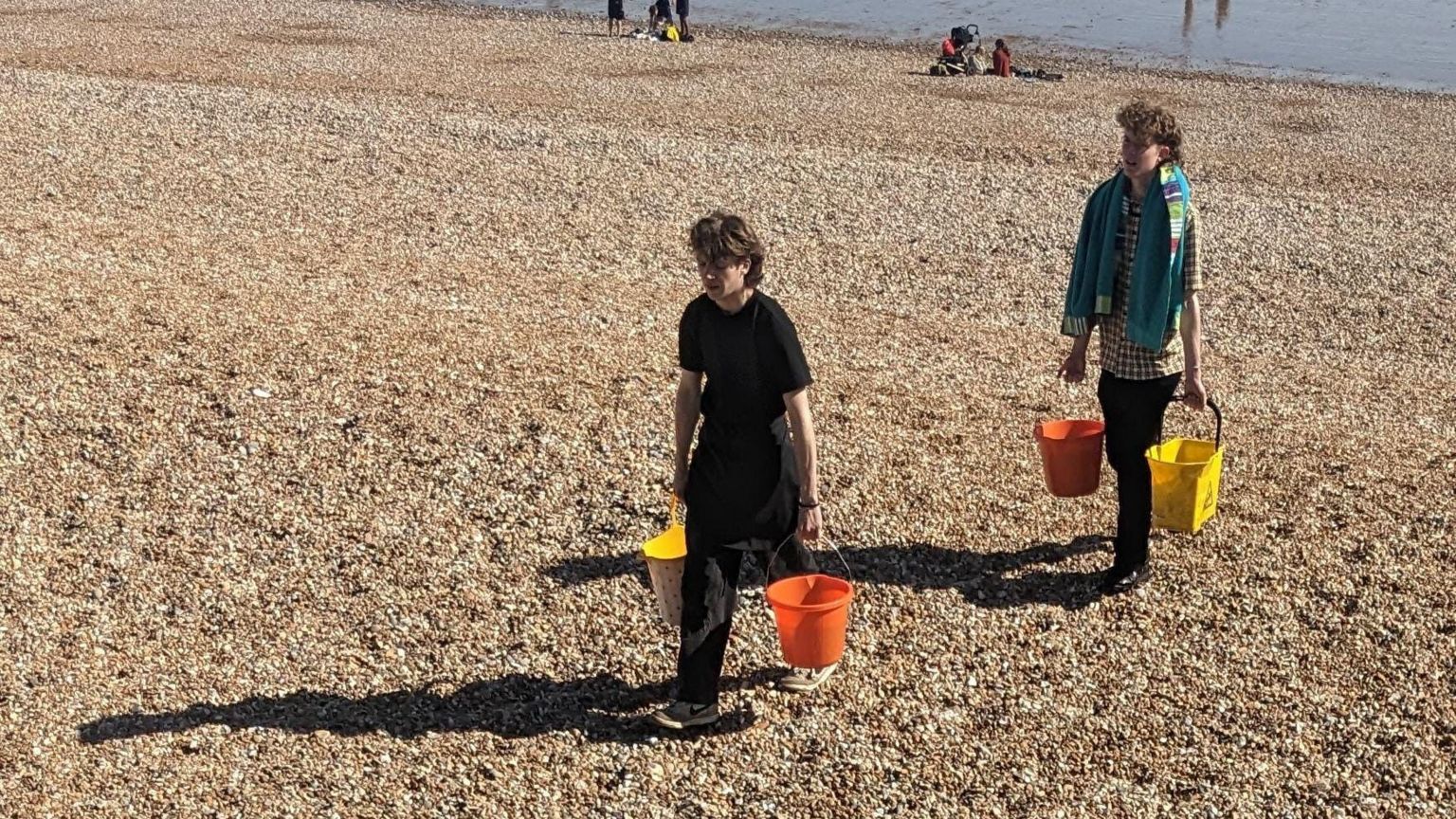 Two young men carry a bucket of sea water in each hand up a stone beach 