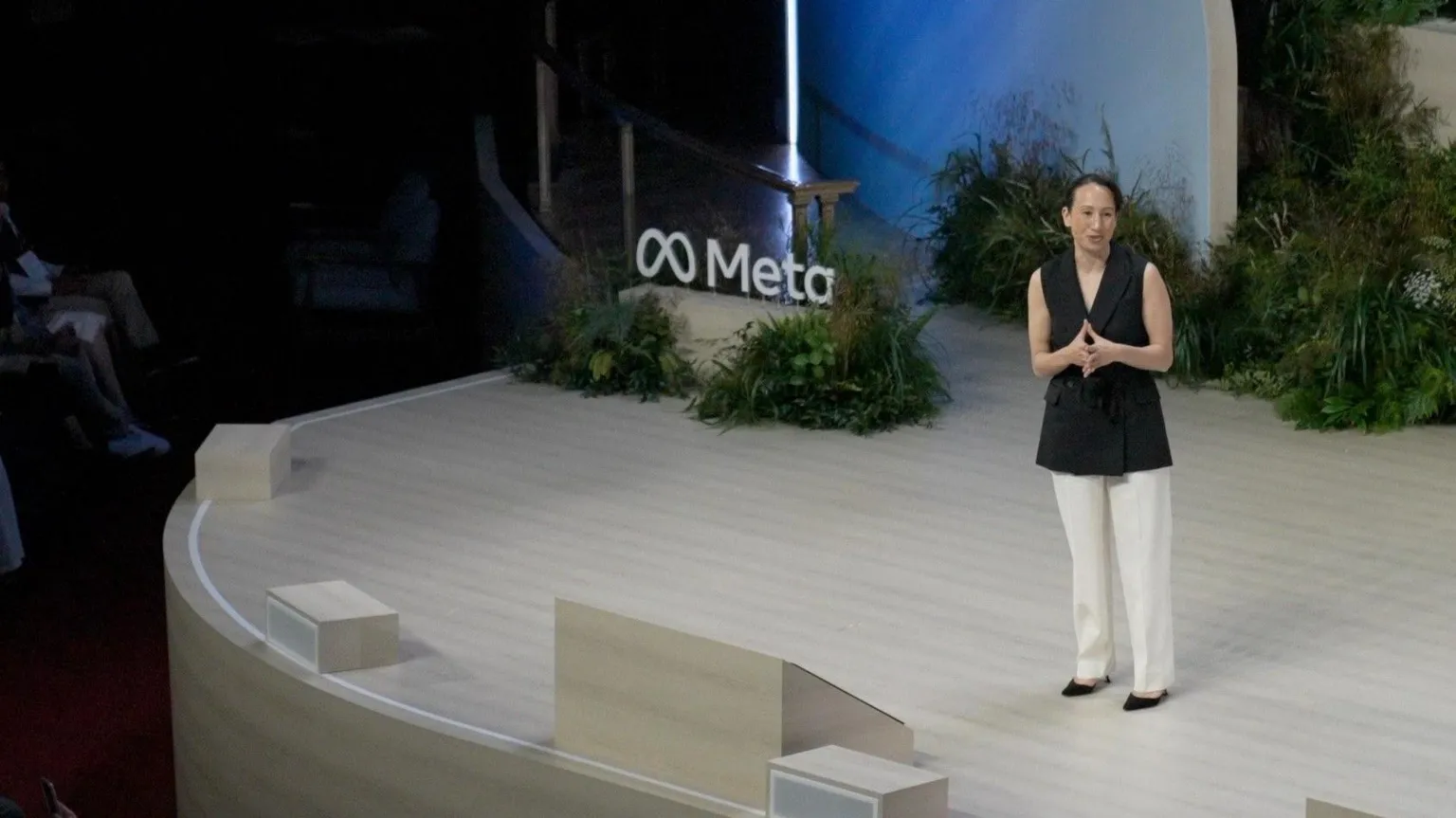 Gleit wears a sleeveless black blazer, white trousers and black shoes as she addresses the audience on-stage at an event in London. A sign bearing Meta's logo is surrounded by foliage behind her on the stage.