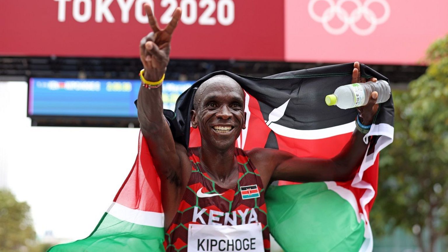 Eliud Kipchoge in Kenya running vest with his name on celebrates his marathon win at the Tokyo Olympics by holding up a Kenya flag behind him and raising two fingers