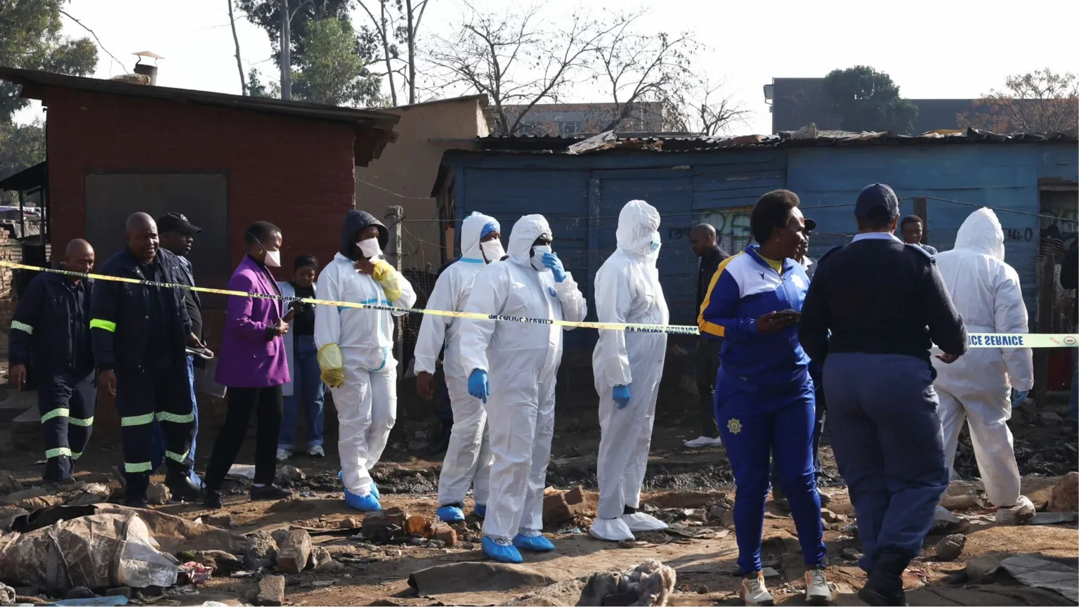 Reuters Forensics team in white PPE suits, masks and blue gloves on the scene where there are police. there is rubbish on the floor and in the background there is police tape in front of some huts