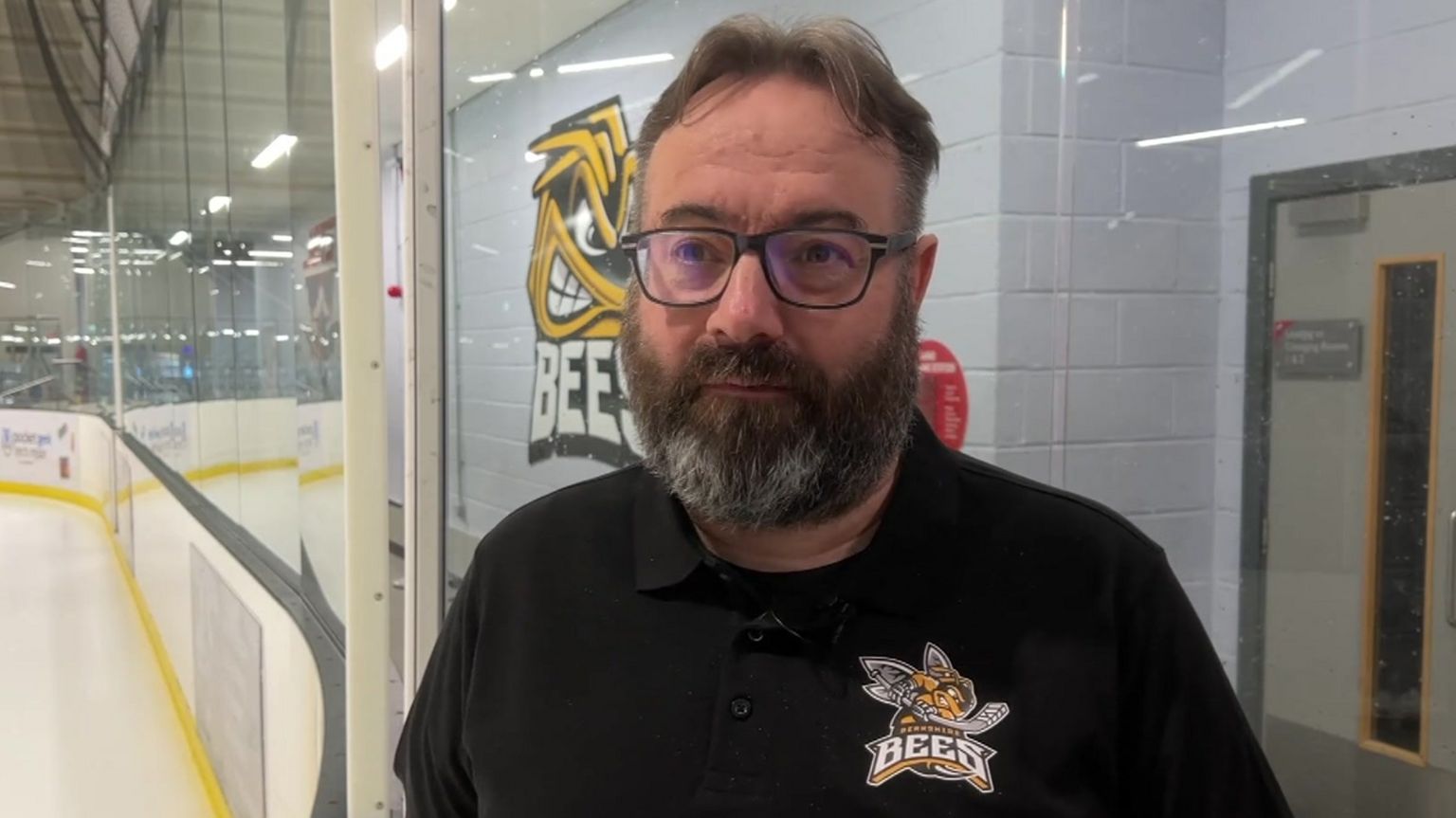 Steve Merry has dark, curtained hair, dark glasses, and a dark beard with flecks of white. He is also wearing a Berkshire Bees branded T-shirt 