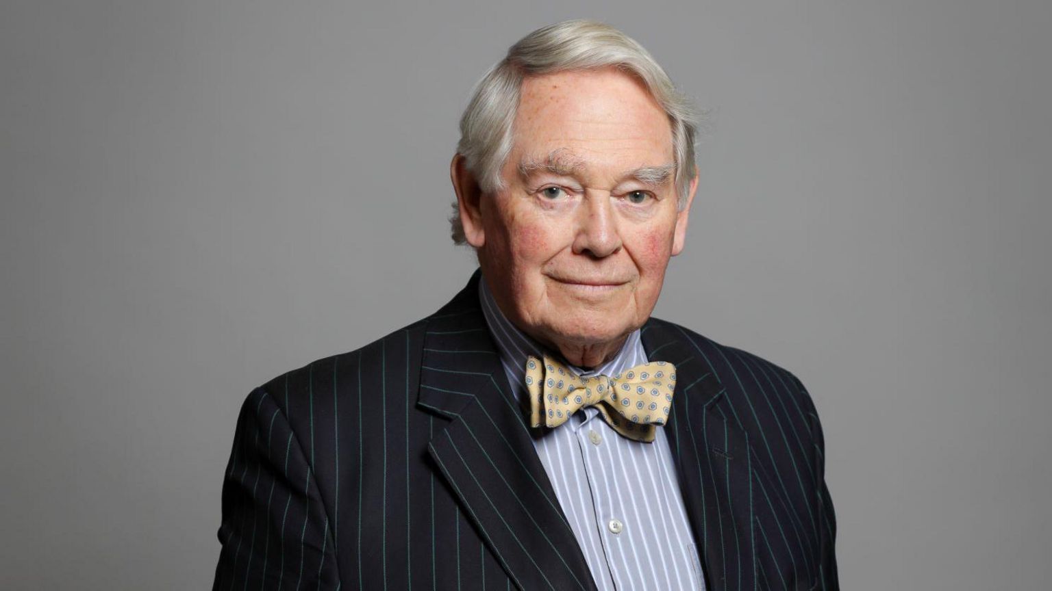 A portrait photo of Lord Naseby wearing a striped shirt, a striped blazer and a yellow and blue bowtie